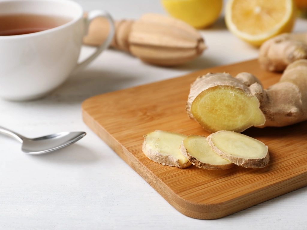 Sliced ginger on a wooden board, next to lemons, a spoon, a lemon squeezer, and a cup of tea