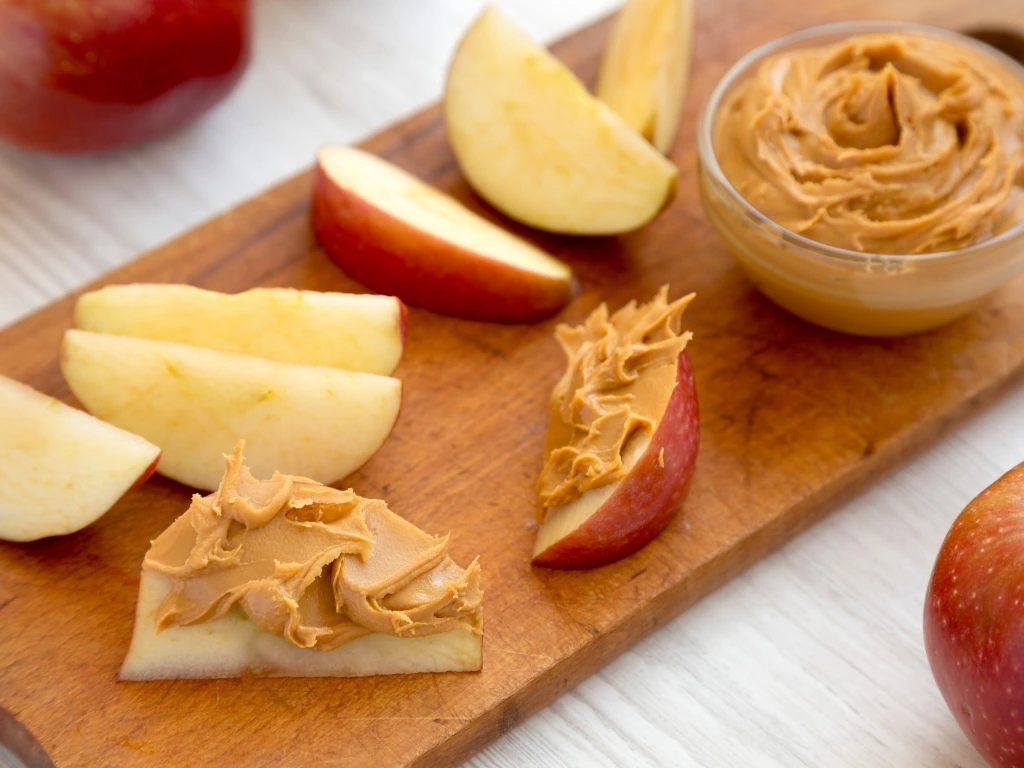 Apple slices with and without nut butter next to a small bowl of nut butter on a wooden board