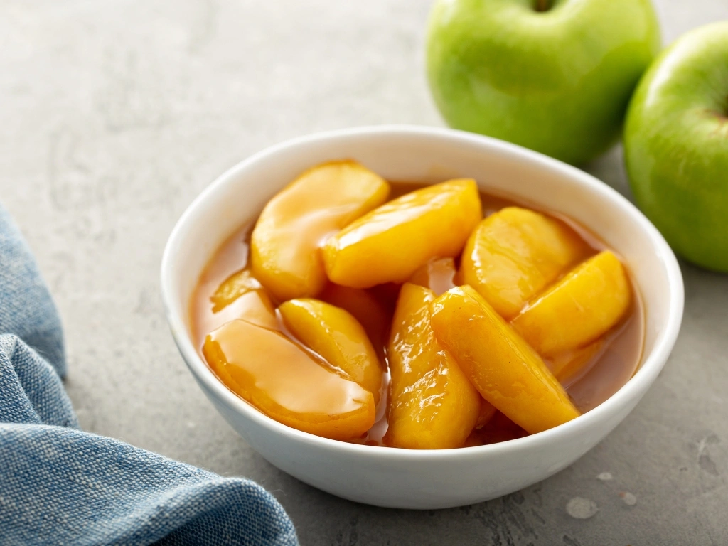 Bowl of sliced peaches in syrup, next to two green apples and a blue tablecloth