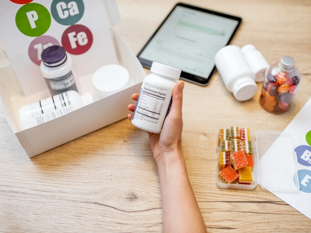 Hand holding a medicine bottle with other bottles, a piece of paper, gummy vitamins, and a tablet on a wooden table