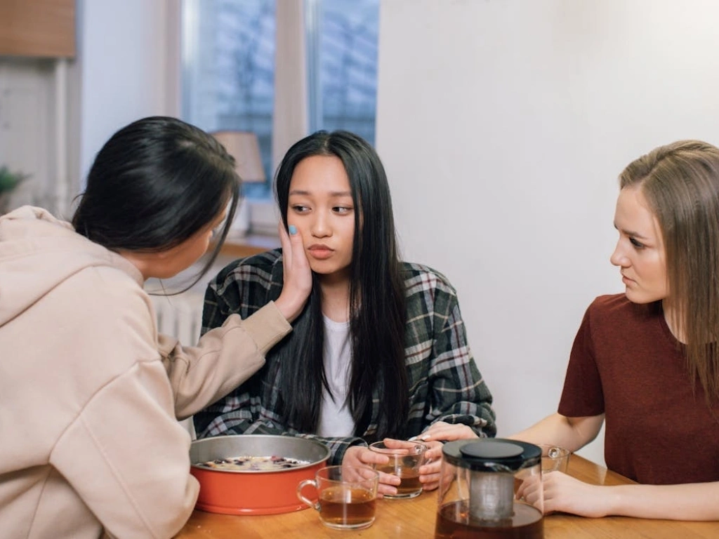 A young woman looking thoughtful or sad while two female friends offer comfort and support at a kitchen table with tea and cake.