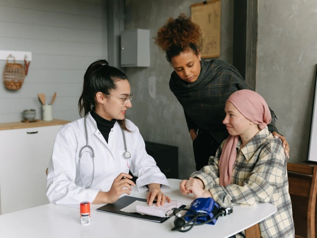 A female doctor in a white coat consulting with a patient wearing a pink headscarf, while a supportive friend or relative stands nearby.