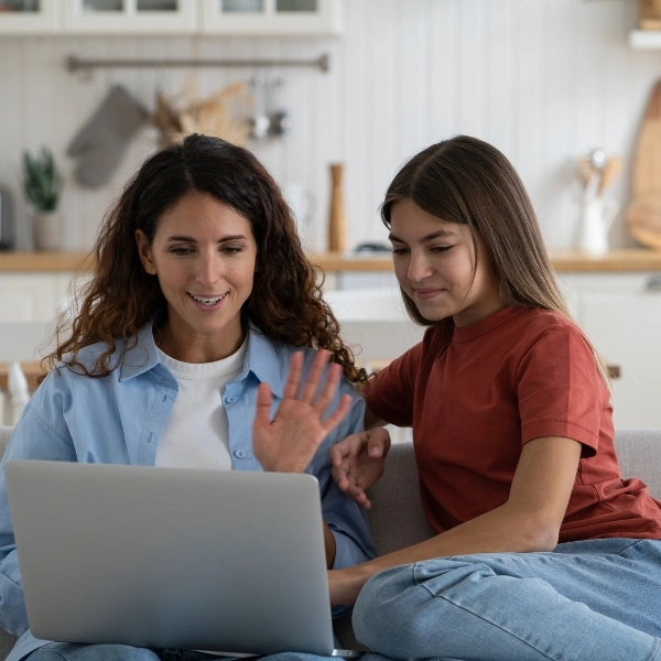 Smiling positive woman and school age girl making video call in laptop sits on couch at home