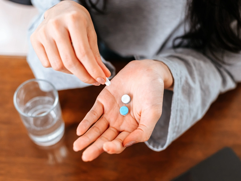 Woman holding a white pill in her fingers above an open palm with two pills, with a glass of water nearby