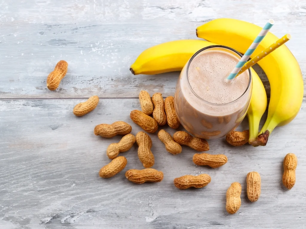 Smoothie with two straws on a wooden table alongside two bananas and a pile of peanuts