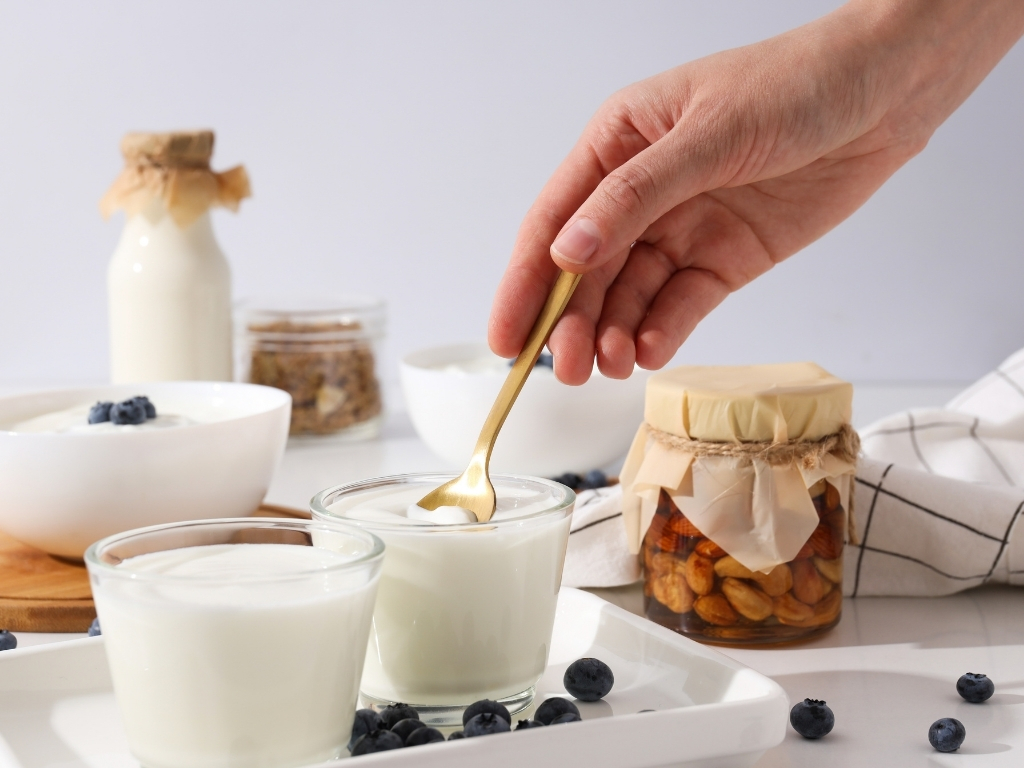 Hand scooping yogurt from a glass with a spoon, with scattered berries and containers of yogurt and nuts on the table