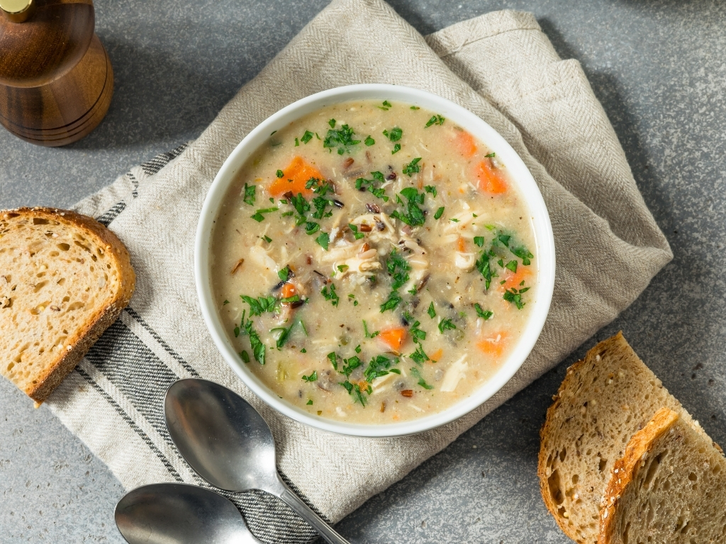  Chicken and vegetable soup in a white bowl, surrounded by slices of bread, a table napkin, a pepper mill, and spoons