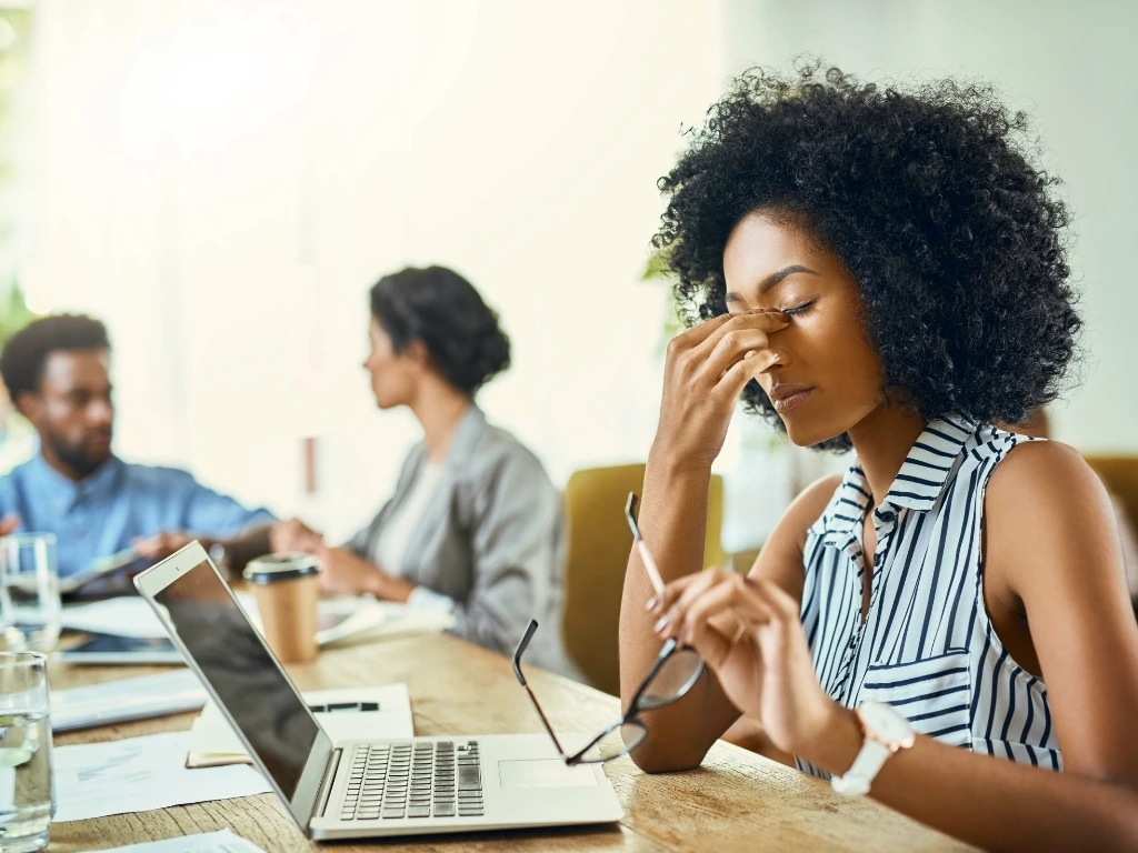 Woman feeling fatigued at work, holding the bridge of her nose with one hand and her eyeglasses with the other, while sitting with colleagues