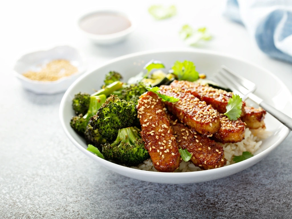White bowl of teriyaki tempeh and charred broccoli served over rice, garnished with sesame seeds