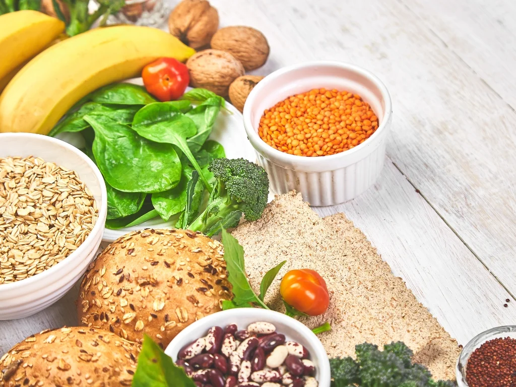 Various vegan foods, including bananas, walnuts, spinach, lentils, broccoli, multi-seed buns, red kidney beans, oats, and seeds, on a white wooden surface