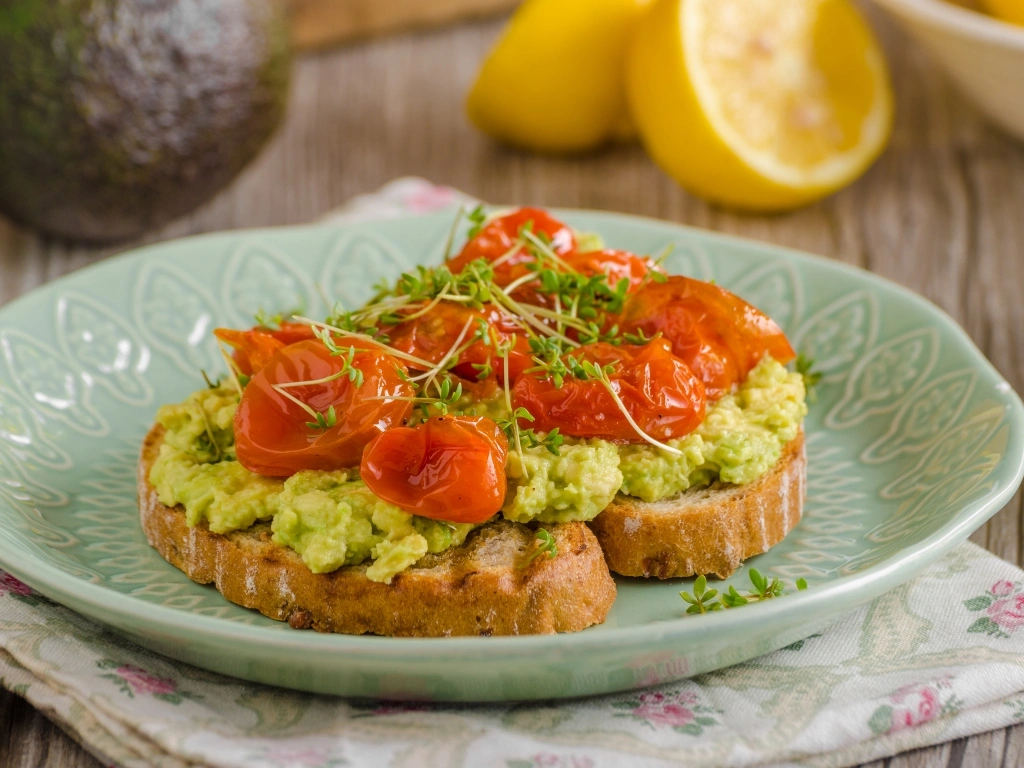 Plate of avocado toast on whole-grain bread topped with roasted cherry tomatoes and fresh microgreens