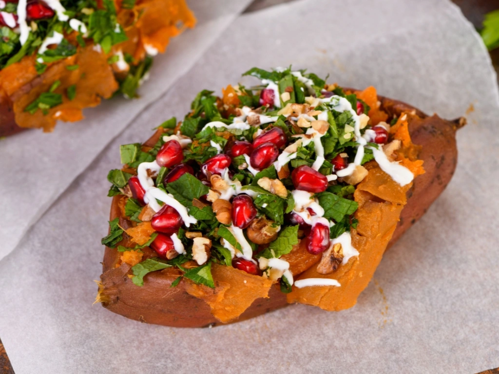 Baked sweet potato stuffed with chopped greens, walnuts, and pomegranate seeds, drizzled with a white sauce