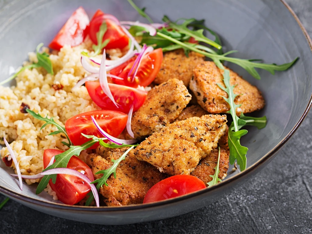 Bowl of cooked quinoa with seasoned protein strips, fresh tomatoes, sliced red onions, and arugula leaves