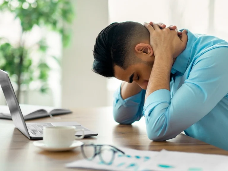 Man feeling fatigued at work, holding his nape with both hands while leaning on a desk with a laptop in front