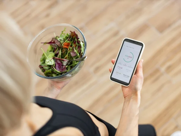 Person using a calorie tracking app with one hand while holding a bowl of salad with the other