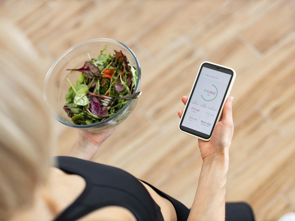 Person using a calorie tracking app with one hand while holding a bowl of salad with the other