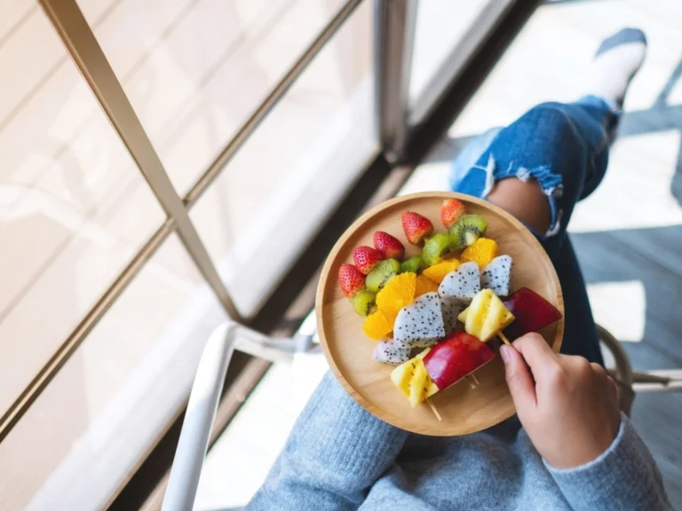Top-down view of a person holding a wooden plate with colorful fruit skewers containing strawberries, kiwi, and dragonfruit