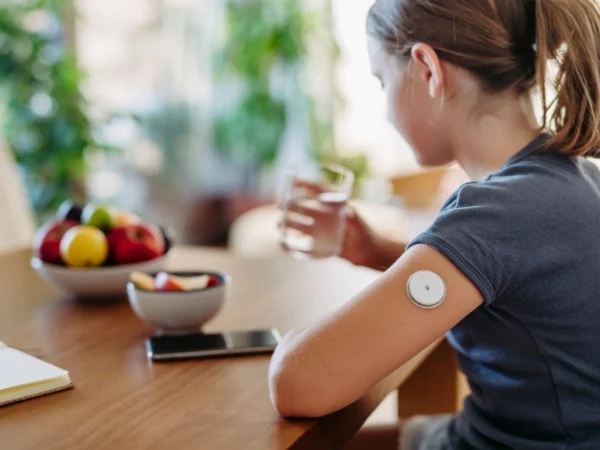 Girl with a continuous glucose monitor sitting by the table holding a glass of water with bowls of sliced and unsliced apples, an open notebook, and a phone in front of her
