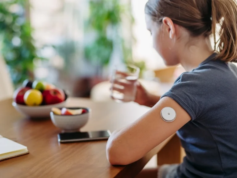Girl with a continuous glucose monitor sitting by the table holding a glass of water with bowls of sliced and unsliced apples, an open notebook, and a phone in front of her