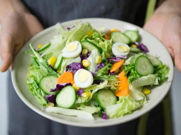 Hands holding a plate of colorful salad with lettuce, cucumbers, boiled eggs, purple cabbage, and corn kernels.
