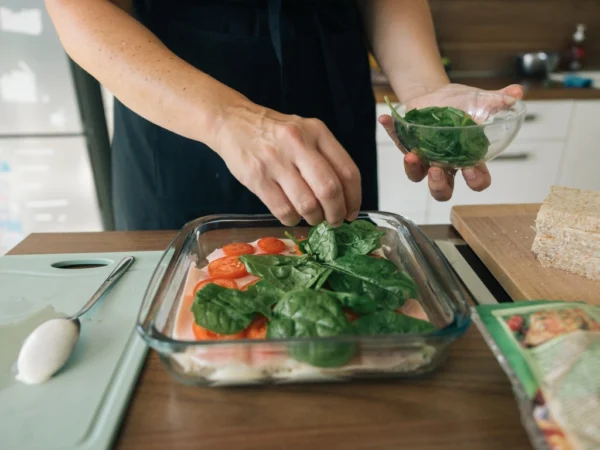 Person adding fresh spinach leaves to a baking dish while preparing a meal in the kitchen