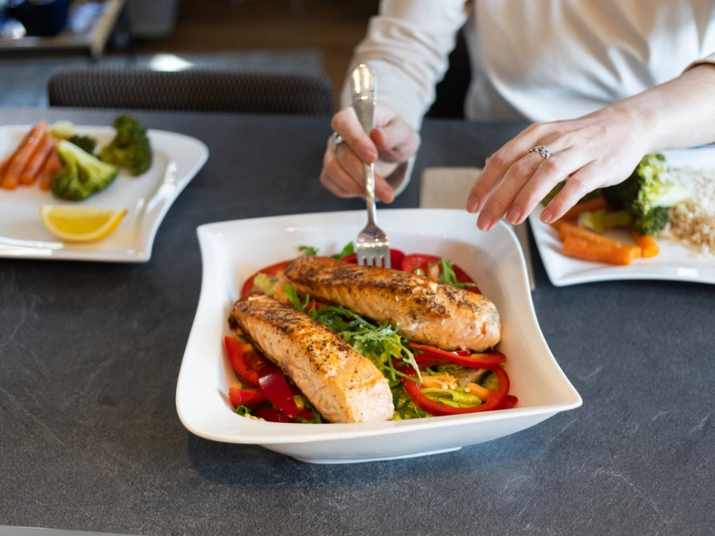 Woman getting a forkful of fish from a plate of two grilled salmon fillets on a bed of spinach and red bell peppers