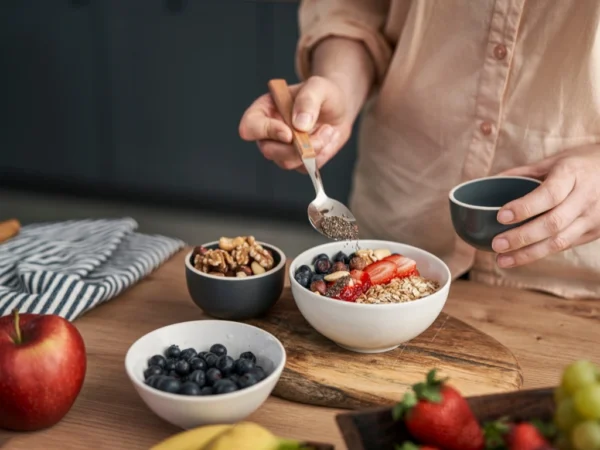 Woman adding a spoonful of seeds into a breakfast bowl with granola, nuts, and fruit, next to bowls of other ingredients