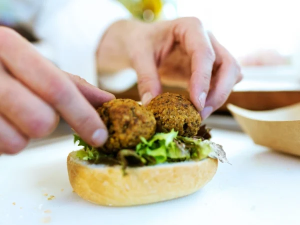 Person preparing falafel meatball sandwich with greens on a white surface