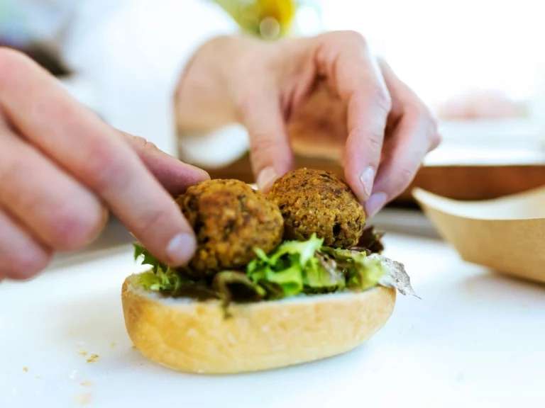 Person preparing falafel meatball sandwich with greens on a white surface