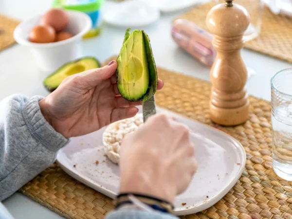 Person slicing a fresh avocado over an English muffin on a plate, surrounded by breakfast items
