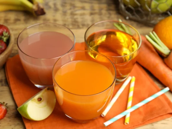 Three glasses filled with juice and herbal tea on a table, next to paper straws, carrots, oranges, apple slices, and strawberries