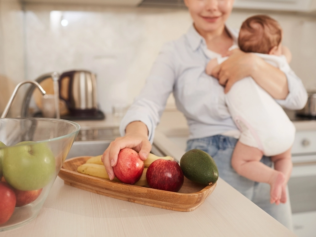 A mother holding her newborn baby while reaching for an apple on the kitchen counter