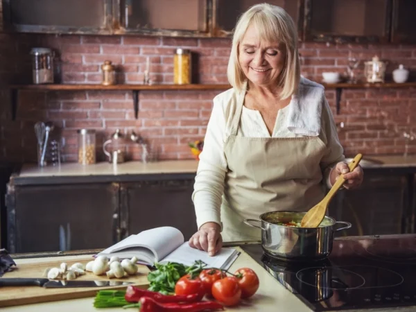 Senior woman in apron is reading a book of recipes, and smiling while cooking in kitchen