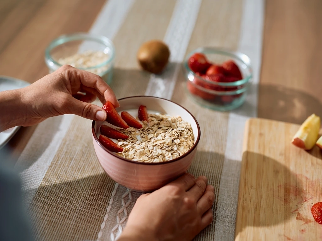 Person adding sliced fruit to a bowl of oatmeal, with various ingredients across the table