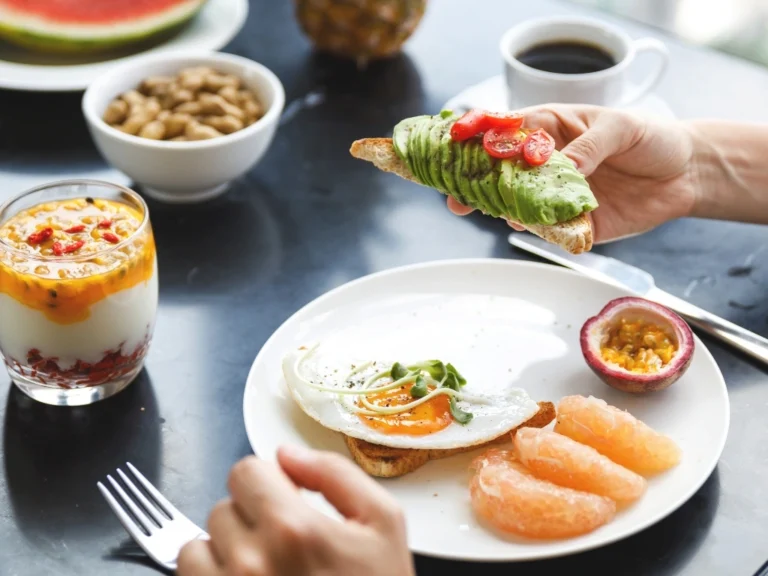 Person holding a piece of toast with avocado, with a spread of healthy foods across the table