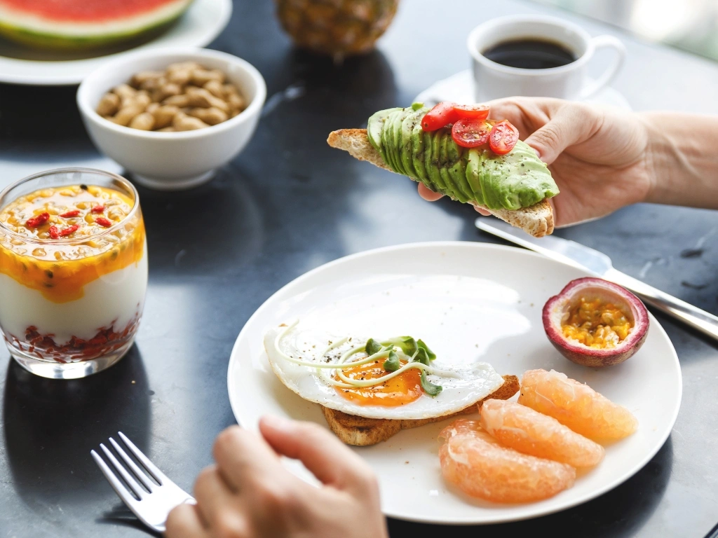 Person holding a piece of toast with avocado, with a spread of healthy foods across the table