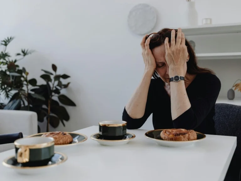 A distressed woman in a black top sits at a dining table with cups of coffee and bread in front of her
