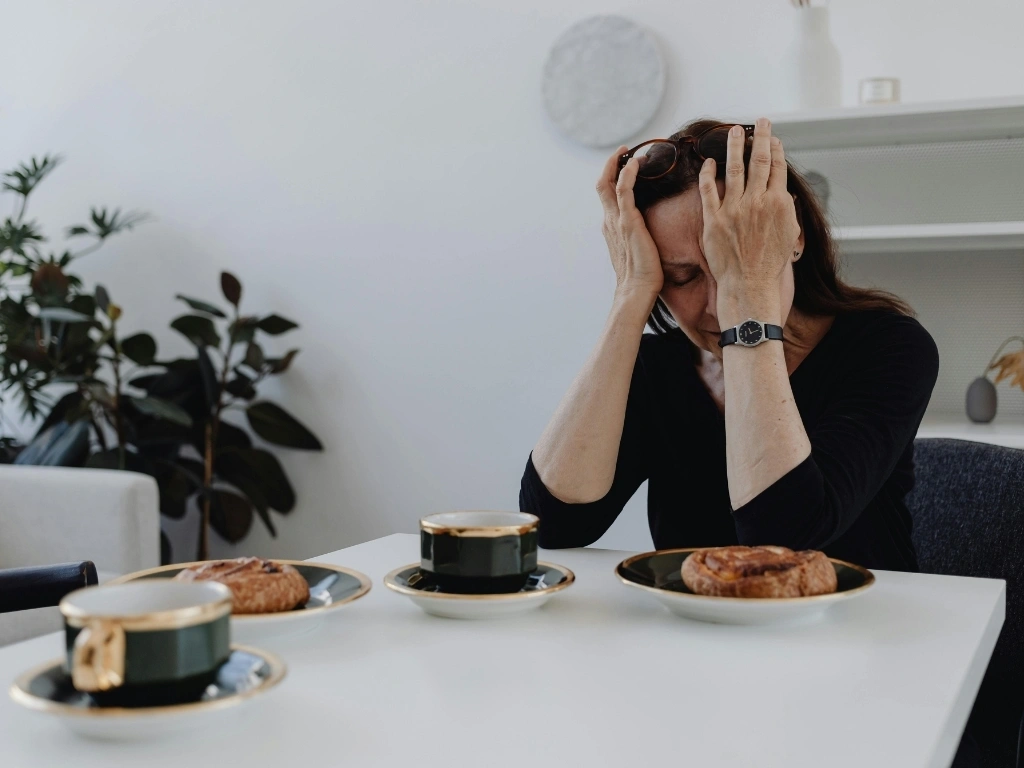A distressed woman in a black top sits at a dining table with cups of coffee and bread in front of her