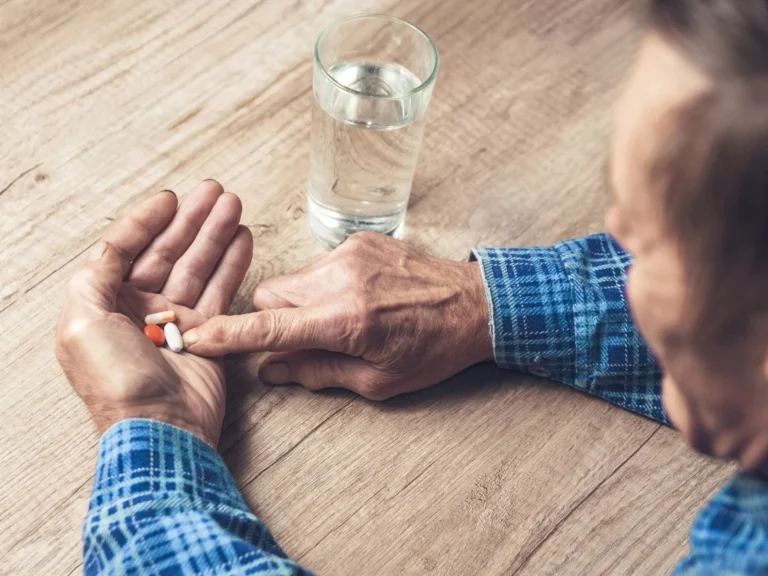 Man seated at a wooden table sorting diabetes medications in his hand, with a glass of water beside him