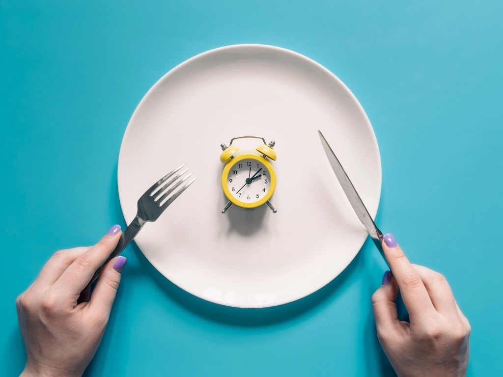 Woman holding utensils, with a small alarm clock served on a plate against a blue surface