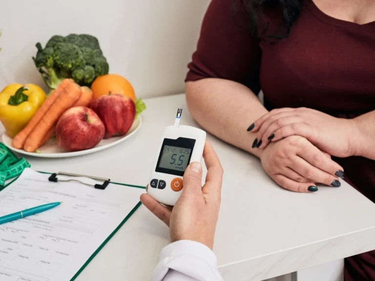 Doctor holding a glucose monitoring device that shows another woman’s blood sugar levels, with a notepad, pen, measuring tape, and a plate of fruits and vegetables on a white table