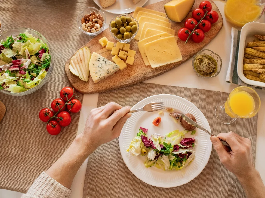 Person eating a salad at a table with a variety of foods including cheese, olives, tomatoes, nuts, and bread