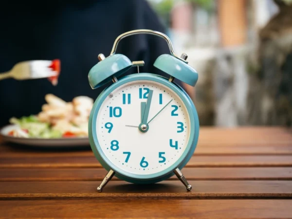 Blue alarm clock on a wooden table in the foreground, with a person eating a meal blurred in the background