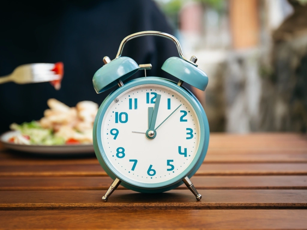 Blue alarm clock on a wooden table in the foreground, with a person eating a meal blurred in the background