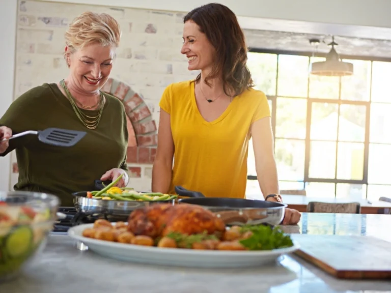 Two women smiling and cooking together in a kitchen with a meal on the counter