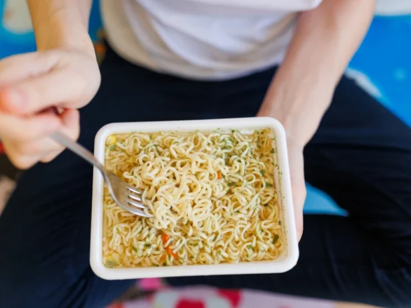 Person holding a square plate full of cooked instant noodles with one hand and a fork with the other