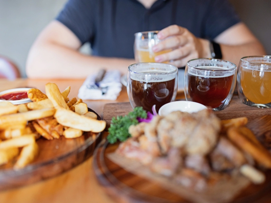 Man seated at a table with different kinds of alcohol, fries, and grilled meat served, holding a glass of beer