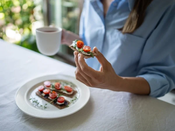 Woman at a table eating crackers with tomato and cheese while drinking coffee