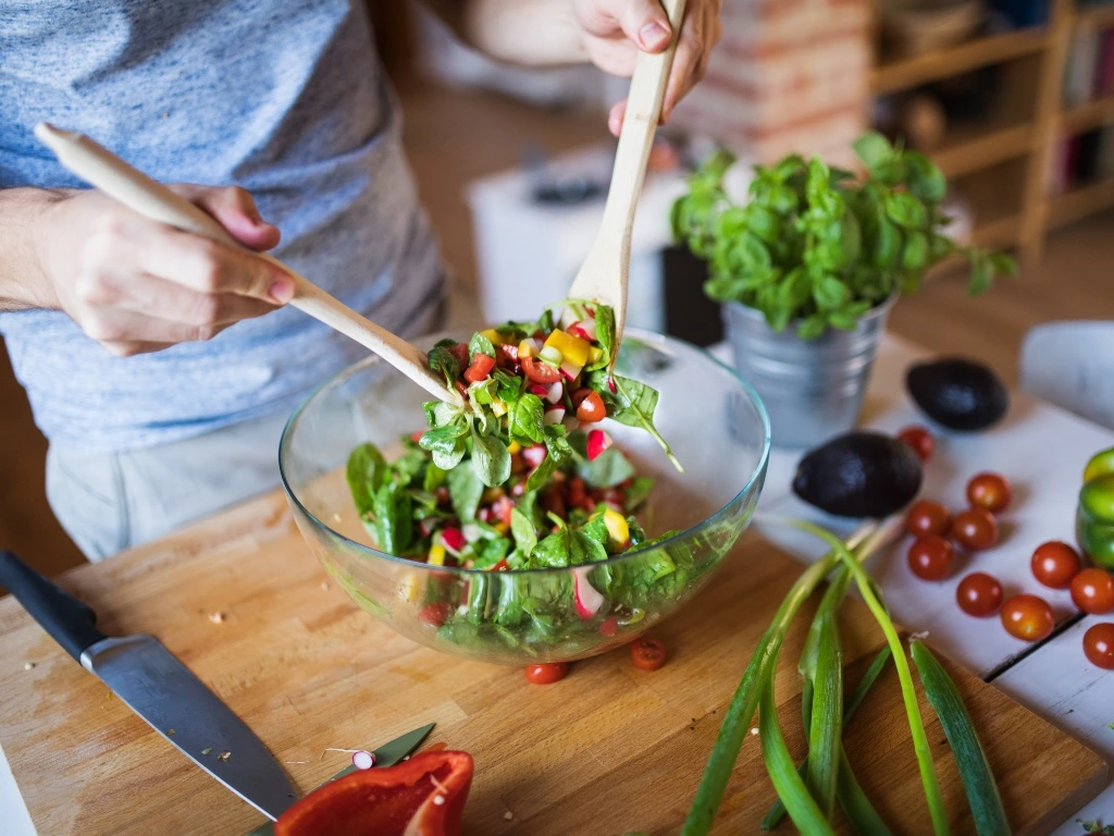Person tossing salad in a bowl using wooden spoons, with different ingredients and a knife laying across the countertop