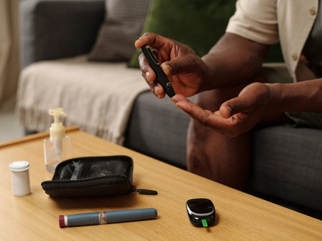 Man seated on a couch using a black lancing device to check his blood sugar, with glucose monitoring supplies arranged on a nearby wooden table